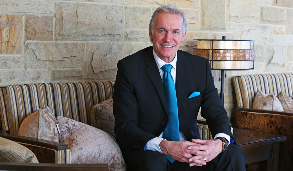 Dr. Hilary Jones seated in a well-lit room, wearing a suit with a blue tie and pocket square, hands folded, against a backdrop of striped cushions and a stone wall.